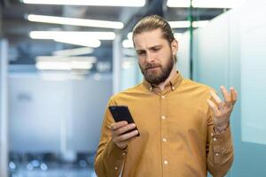 A man with a confused expression holds a smartphone, seeming frustrated or puzzled, in a bright, modern setting. photo