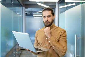 Confident young professional holding a laptop, contemplating ideas and working in an office. The image expresses focus, problem-solving, and engagement in a professional environment. photo