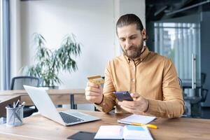 A professional man purchases online using a credit card and smartphone in an office setting, displaying modern technology usage for convenient transactions at the workplace. photo