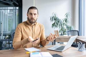 A thoughtful man seated at a desk with a credit card and smartphone, appearing puzzled, possibly about an online transaction or banking issue. photo