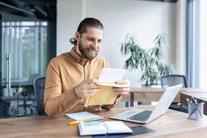 A businessman sits at his desk, smiling as he opens an envelope, amidst a professional environment. Perfect for concepts related to business, communication, office ambiance, and workplace joy. photo