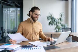 Successful individual concentrated on reviewing paperwork while using a laptop in a bright office. He appears professional, engaged, and content as he manages work efficiently. photo