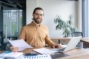 Smiling office worker sitting at desk with laptop, reviewing documents, and analyzing reports. The workspace exudes professionalism and productivity photo