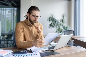A professional is sitting at a desk in an office setting, analyzing papers and using a laptop. The workplace is equipped with modern elements, fostering concentration and productivity. photo