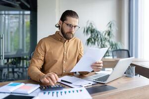 A professional male working at a desk, examining papers, and using a laptop and calculator. Depicts productivity, focus, and office work in a business or financial setting. photo