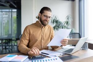 A focused professional reviews paperwork while working on financial or business tasks at a desk with a laptop and calculator, suggesting determination, productivity, and a modern office environment. photo