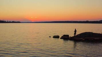 Fisherman at the evening river. Silhouette of a man fishing on the stone in water on the beautiful nature background at sunset. video