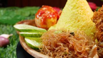 Close Up cone shaped yellow rice served with crispy fried chicken, red sambal, sambal egg, fried vermicelli, fresh cucumber slices, along with red onions, garlic, and peppercorns in small bowls. video