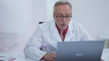 Medical worker having distance call. Senior doctor in white gown sitting in front of computer and talking to a client in medical center. Online consultation. video