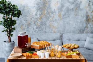 There is a wellarranged table featuring a diverse array of delicious food and refreshing drinks set up in a cozy living room space, inviting everyone to gather and enjoy photo