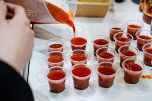 A person is pouring a rich and flavorful sauce into several plastic cups that are arranged neatly on a table, ready for serving or further preparation. It looks delicious photo