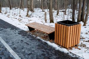 A sturdy wooden bench, complemented by a trash can, sits quietly on the side of a snowcovered road, which together create a serene winter scene filled with immense beauty and tranquility photo