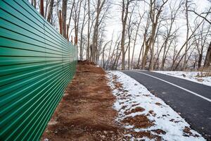 A vibrant green corrugated fence runs alongside a winter walking path set in a tranquil leafless forest, creating a picturesque scene for anyone enjoying the natural wilderness photo