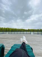 A person is comfortably lying back on a bench with their feet elevated, enjoying the view of the sky above, surrounded by fluffy clouds and the lush green grass all around them photo