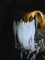 A person is carefully walking through a natural cave that features stunning and unique ice formations hanging from the ceiling and surrounding the paths, creating a breathtaking scene photo