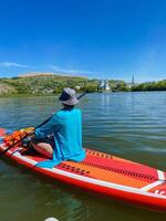 A woman is comfortably sitting on a paddle board that is floating on the surface of a serene lake, surrounded by picturesque scenery and enjoying the beautiful day outdoors photo