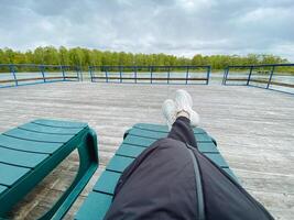 A person is comfortably laying back on a chair positioned on a dock, enjoying a serene view with their feet propped up, basking in the daylight and relaxation, surrounded by nature photo