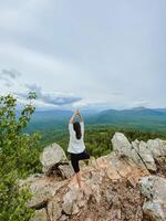 A woman is diligently practicing her yoga poses atop a majestic and rocky mountain, surrounded by the breathtaking views that extend to the sky above and the clouds nearby photo