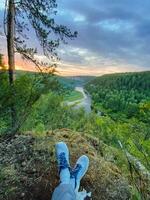A solitary individual is sitting perched on the edge of a tall cliff while taking in the breathtaking view of a winding river far below, surrounded by the beauty of nature photo