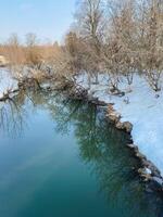 A beautiful river flows gracefully, adorned with snow on its tranquil shores, while trees stand tall in the background, creating a serene and picturesque natural landscape photo