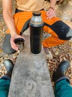 A person sits comfortably on a wooden bench, relishing a steaming hot drink poured from a thermos and sipping coffee from a cup, while surrounded by the breathtaking beauty of nature photo