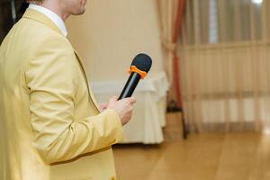 A man in a yellow jacket is standing and holding a microphone, probably giving a speech at a formal event. The setting includes a wooden flooring and a public address system photo