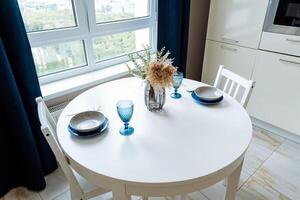 The kitchen is elegantly set with a white table adorned with plates, glasses, and a vase of flowers. Its interior includes a window, chair, and plant, reflecting a tasteful design photo