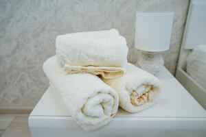 A sleek white table, situated next to a warm and cozy lamp, holds three neatly rolled towels, which enhances the clean and inviting atmosphere, promoting both comfort and stylish elegance photo