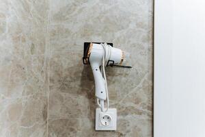 A hair dryer is currently hanging securely on a wall right next to an electrical socket, making it conveniently accessible for use whenever it is needed for drying hair or styling photo