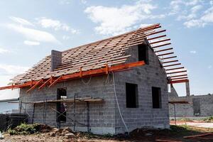 A brick house is currently under construction, featuring a wooden roof that is being carefully placed. The project site is surrounded by various plants and an expansive land lot photo