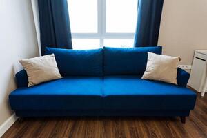 A cozy living room features an azure couch, white pillows, elegant furniture, hardwood flooring, and natural light, creating a stylish and comfortable space for relaxation and socializing photo