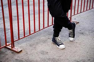 An individual is standing by a bright red fence, casually crossing their legs in an eyecatching pose that captures the attention of those nearby, enhancing and elevating the overall scene photo