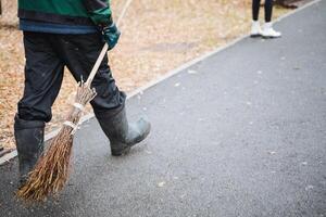 A man is leisurely walking down the street while carrying a broom in his hands, possibly to clean up any debris he might find along the way. This situation could be seen on an urban road photo