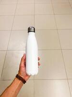 A person is standing, holding a white plastic water bottle with their fingers wrapped around it. It is clear the bottle contains liquid. The scene is likely indoors on a floor surface photo