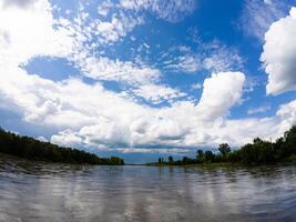 A serene and tranquil lake, beautifully surrounded by vibrant green trees, mirrors fluffy white clouds floating in a bright blue sky, creating a stunning and picturesque natural landscape photo