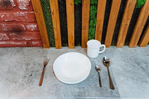On a charming table positioned in front of a rustic wooden fence, there lies a beautifully arranged dining set consisting of a plate, a cup, spoons, and a fork, creating a delightful scene photo