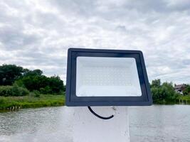 A powerful flood light is positioned on top of a tall white pole, which stands next to a serene body of water, likely a lake that reflects the surrounding landscape beautifully photo