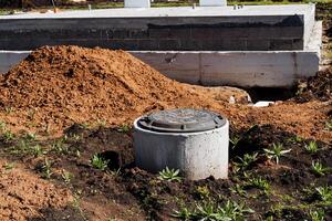 Construction in progress at site with concrete manhole cover and soil piles under sunny sky. Green plants surround area as workers install piping for infrastructure project photo