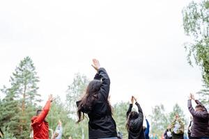A joyful group of people gathers in a circle outdoors, engaging in a leisure activity under the sky surrounded by trees, nature, and green grass. They raise their arms in excitement photo