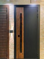 There is a striking black door that features a wooden handle right next to a sturdy brick wall, creating a contrasting yet harmonious visual effect between the surfaces around it photo