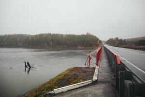 An asphalt bridge over a serene lake, with trees under a cloudy sky photo