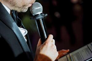 Man in formal wear gestures with microphone while reading paper photo