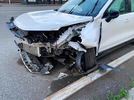 A white car with a damaged front end is parked on the side of the road, possibly due to a collision. The wheel, tire, hood, and grille seem affected, indicating significant damage photo