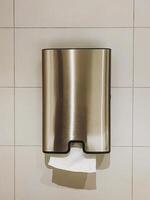 A shiny stainless steel paper towel dispenser, designed in a sleek rectangular shape, is hanging on a tiled wall that adds a touch of modernity to the restroom or kitchen area photo