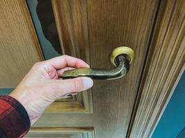 A hand clasping a vintage brass door handle positioned on an elegantly designed wooden door creates a charming and inviting entrance photo