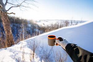 Tourist utensils, a mug for tea, a glass for hot drinks are held in the hand of a man against the background of a winter forest. photo