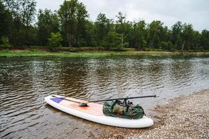 The SUP board is parked to the shore, tourist equipment for rafting on a mountain river, a dry bag for things is fixed on a board, an inflatable SUP. photo