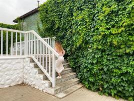 A woman is elegantly climbing a staircase next to a wall adorned with ivy vines, creating a picturesque scene of nature and architecture. The setting includes plants, stairs, and a building photo