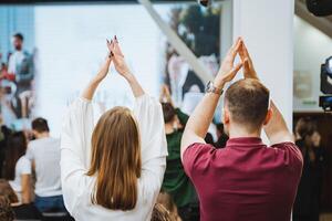 A man and woman are gesturing in front of a large screen at an event photo