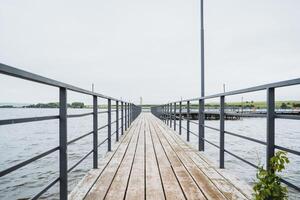 A wooden pier with a metal railing extends over calm water beneath a clear sky, forming a peaceful scene by the lake. It provides a serene spot for relaxation and contemplation photo